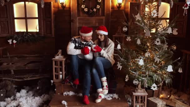 Vriend en vriendin in Santa hoeden zittend op veranda naast de kerstboom. - Video