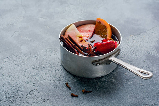 Mulled wine hot drink with citrus, apple, pomegranate and spices in aluminum casserole with vintage christmas tree toys and Fir branch on concrete background. - Photo, image