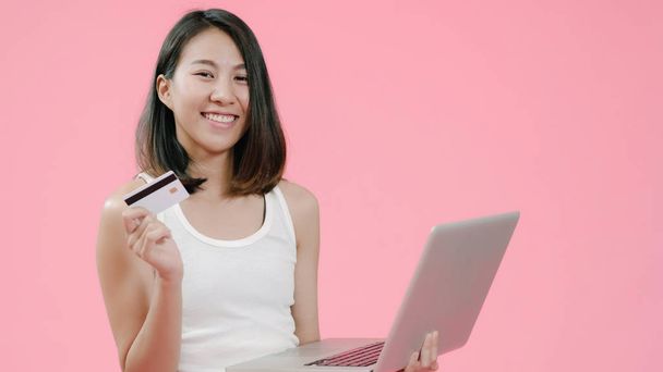 junge asiatische Frau mit auf dem Laptop Online-Einkauf per Kreditkarte in lässiger Kleidung und Blick auf den Computer über rosa Hintergrund Studioaufnahme. glücklich lächelnd liebenswert fröhlich Frau freut sich über Erfolg. - Foto, Bild