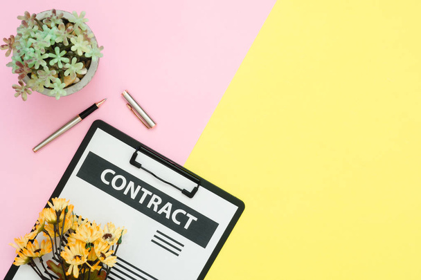 Flat lay design of office desk working space  - Top view of a contract document and a pen with yellow flower and tree on pink and yellow pastel color with copy space. Pastel working space background. - Photo, image
