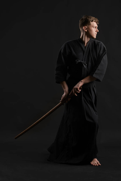 Kendo guru vestindo em um kimono japonês tradicional está praticando arte marcial com a espada de bambu de xadrez contra um fundo de estúdio preto. - Foto, Imagem