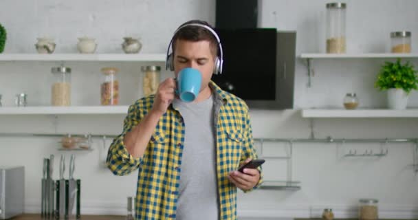 Young man enjoys morning time, drinking tea and listening music in headsets - Záběry, video