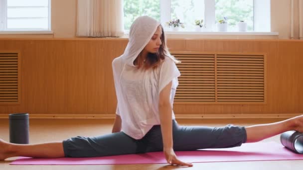 Giovane donna magra seduta sul tappetino da yoga in split - con supporto ausiliario sotto un piede - appoggiata in avanti
 - Filmati, video