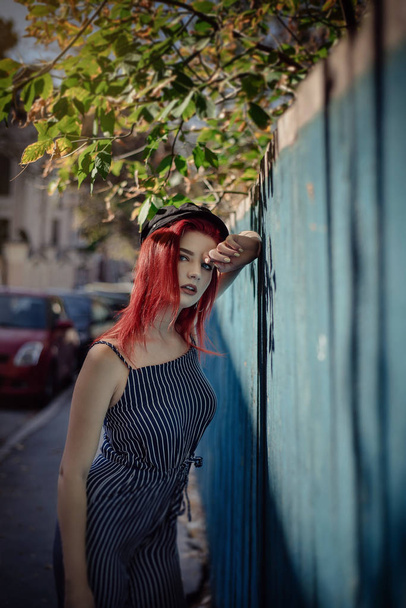 La mujer joven a la moda en la ropa en la raya y el cabello rojo, posando sobre el fondo azul en la calle
. - Foto, imagen