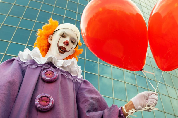 Street makeup on the face of a teenager. A boy in the image of a clown with sharp teeth holds in his hand two red balloons against the background of a modern building made of glass. Halloween. - Photo, Image