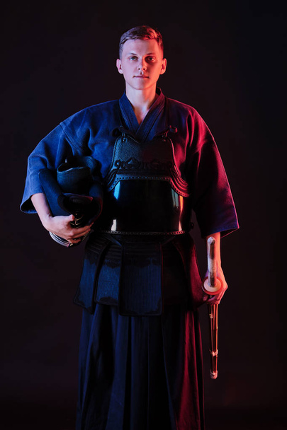 Kendo-Kämpfer in einer Rüstung, traditioneller Kimono hält seinen Helm und sein Shinai-Bambusschwert, während er auf schwarzem Hintergrund posiert. Nahaufnahme. - Foto, Bild