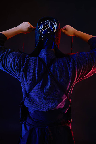 Close up shot, Kendo fighter wearing in an armor and traditional kimono is tying the lacing on his helmet standing back against a black background. - Fotografie, Obrázek