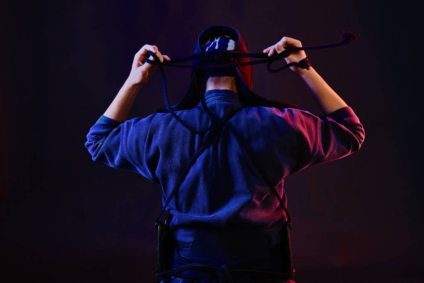 Cerrado el tiro, el luchador de Kendo con armadura y kimono tradicional está atando la laca de su casco de pie contra un fondo negro.. - Foto, imagen