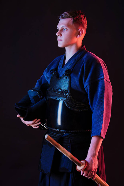 Боец кендо в доспехах, традиционном кимоно держит в руках шлем и меч синай-бу, позируя на черном фоне. Закрыть. - Фото, изображение