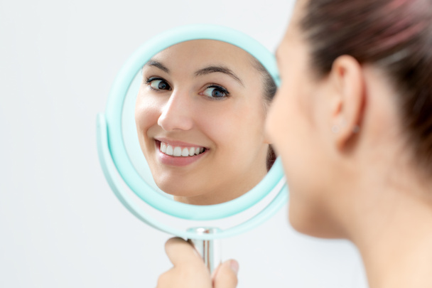 Закрыть портрет молодой женщины, смотрящей в зеркало на себя. Девушка со здоровыми белыми зубами держит ручное зеркало
. - Фото, изображение