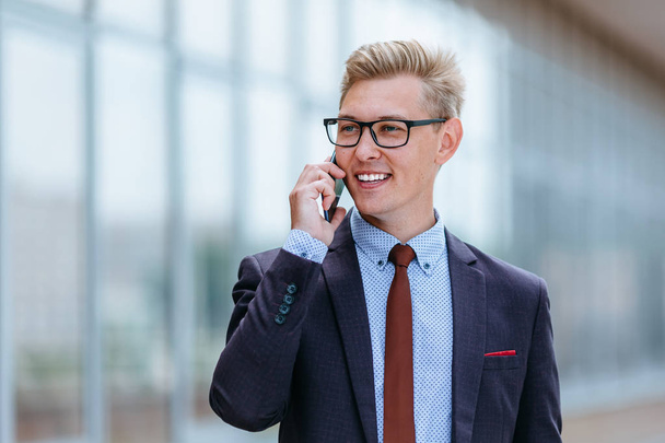 Young handsome Caucasian businessman in glasses talking on mobile phone in front of office building. Male executive manager in trendy clothes using smartphone outdoors. Modern technology concept - Fotoğraf, Görsel