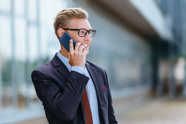 Young handsome Caucasian businessman in glasses talking on mobile phone in front of office building. Male executive manager in trendy clothes using smartphone outdoors. Modern technology concept - Фото, зображення
