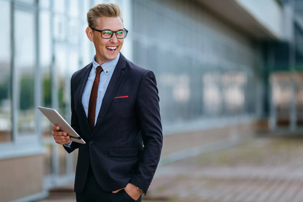 Elegant smiling young businessman using his PC tablet. Portrait of a Male executive manager in trendy clothes with tablet pc in front of office building. Modern technology concept - Zdjęcie, obraz