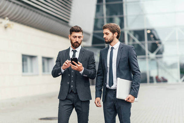 Two attractive smiling young businessmen wearing suits walking outdoors at the city streets, using mobile phone - Фото, зображення