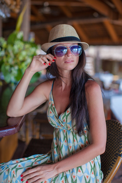 Beautiful brunette woman in long dress and hat relaxing near bar in exotic resort - Foto, Imagen