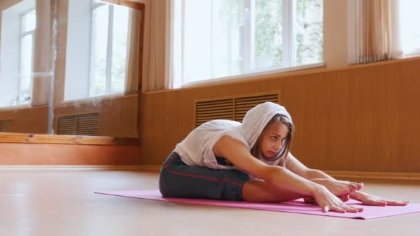 Молодая стройная женщина в белой толстовке сидит на коврике для йоги и наклоняется вперед - наклоняясь и касаясь земли над ногами
 - Кадры, видео