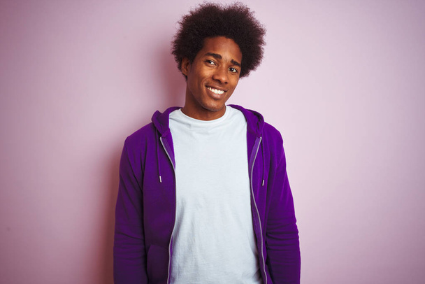 孤立したピンクの背景の上に立つ紫色のスウェットシャツを着た若いアフリカ系アメリカ人男性が、横を見つめて考え直して微笑んでいる. - 写真・画像