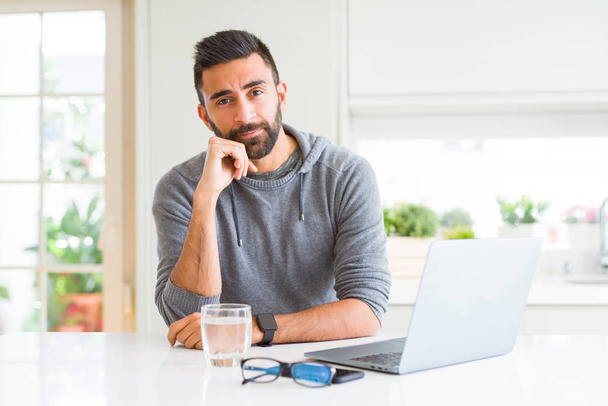 Hombre hispano guapo trabajando con computadora portátil con la mano en la barbilla pensando en la pregunta, la expresión pensativa. Sonriendo con cara pensativa. Concepto de duda
. - Foto, imagen