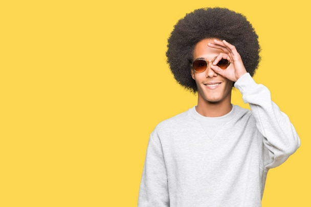 Young african american man with afro hair wearing sunglasses doing ok gesture with hand smiling, eye looking through fingers with happy face. - Fotó, kép