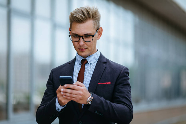 Happy businessman smiling as he reads a text message standing outside the office. Male executive manager in trendy clothes using smartphone outdoors. Modern technology concept - 写真・画像