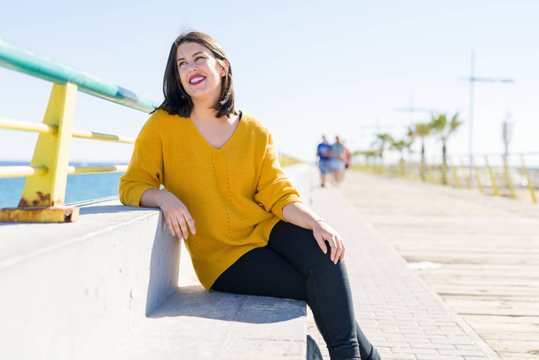 Mulher bonita sentada em um passeio junto ao mar sorrindo alegre desfrutando de luz solar relaxante no dia ensolarado
 - Foto, Imagem