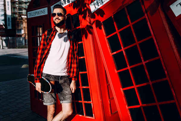 Stylish man with a beard, hipster holds a skateboard in his hands and poses in front of a red telephone box - Фото, зображення