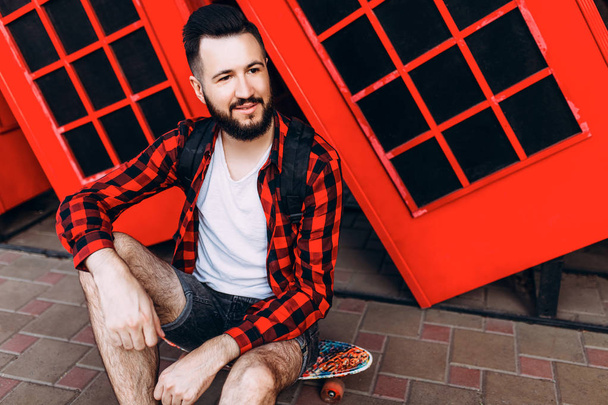 Stylish man with a beard, a man holding a skateboard in his hands and posing in the city in front of a red telephone box - Foto, immagini