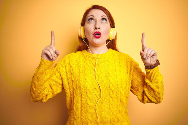 Mladá rudovlasá žena naslouchající hudbě pomocí sluchátek přes žlutý izolovaný podklad užaslý a překvapený pohledem a ukazujícími prsty a zdviženými pažemi. - Fotografie, Obrázek