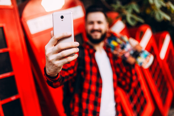 man with a beard in sunglasses, a hipster dressed in a plaid shirt with a skateboard, takes a selfie on a smartphone - 写真・画像