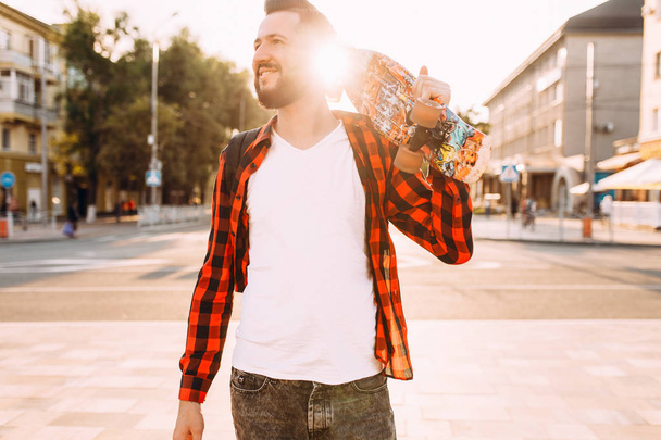 guy with a beard, a hipster walks around the city with a skateboard in his hands. - Фото, изображение