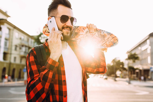 あごひげを生やした男は、スケートボードを持って街を歩き回り、スマートフォンで話す - 写真・画像