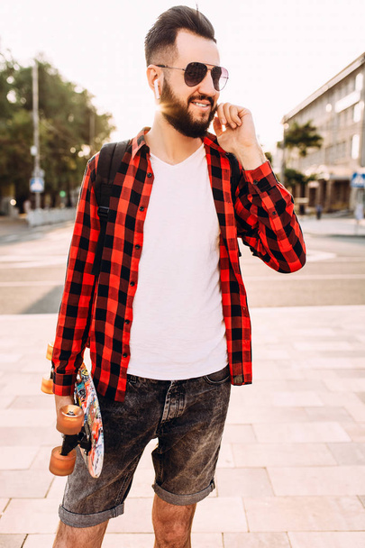 Stylish guy with a beard in wireless headphones walks with a skateboard around the city - Фото, изображение