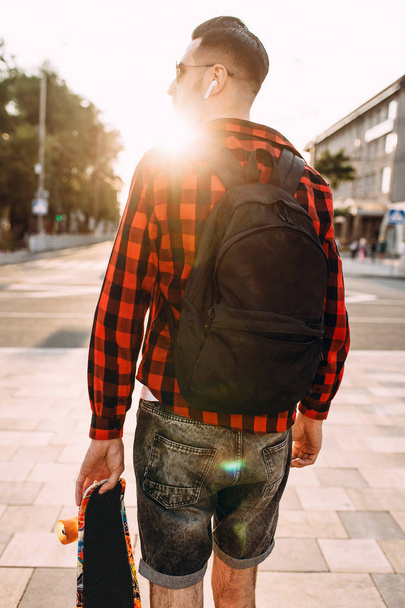 Stylish guy with a beard in wireless headphones walks with a skateboard around the city - Фото, зображення