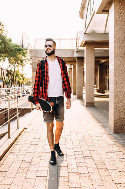 Stylish guy with a beard in wireless headphones walks with a skateboard around the city - 写真・画像