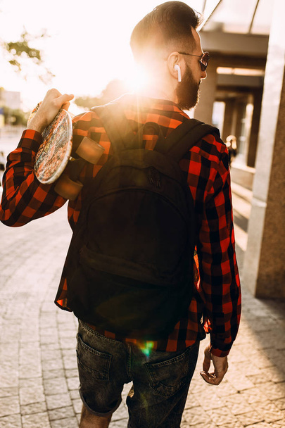 Stylish guy with a beard in wireless headphones walks around the city with a skateboard - Fotó, kép