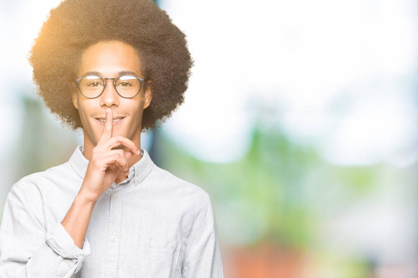 Joven afroamericano con cabello afro usando gafas pidiendo estar callado con el dedo en los labios. Silencio y concepto secreto
. - Foto, Imagen