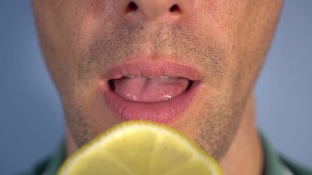 A young man with bristles puts in his mouth and chews a large piece of sour lemon. In a green polo shirt on a blue background. The fight against vitamin deficiency and colds - Imágenes, Vídeo