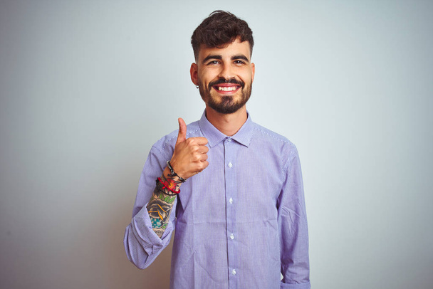Nuori mies tatuointi yllään violetti paita seisoo yli eristetty valkoinen tausta tekee onnellinen peukut ylös ele käsin. Hyväksymällä ilme katsomalla kameraa osoittaa menestystä
. - Valokuva, kuva