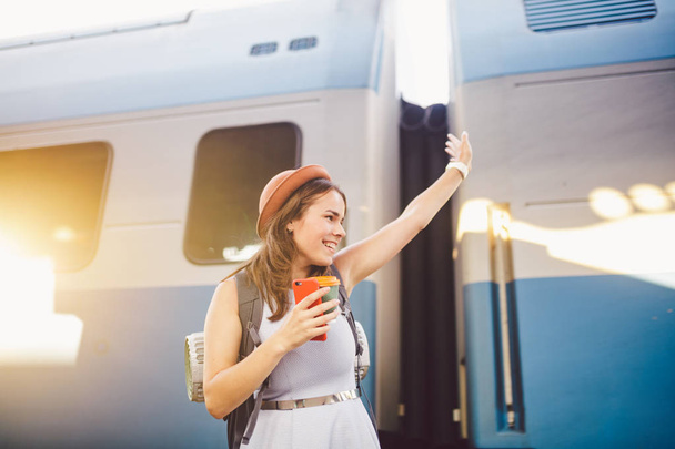 рюкзак путешественник женщина машет рукой на вокзале платформы летние каникулы концепции путешествия. Женское приветствие и удовольствие от поездки на поезде, концепция путешествия. Тематический транспорт и путешествия
 - Фото, изображение
