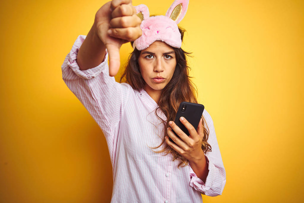 Mujer joven con pijama y máscara usando teléfono inteligente sobre fondo aislado amarillo con la cara enojada, signo negativo que muestra aversión con pulgares hacia abajo, concepto de rechazo
 - Foto, Imagen