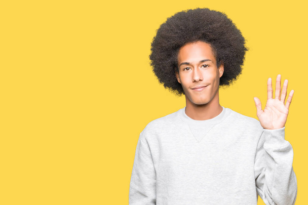 Young african american man with afro hair wearing sporty sweatshirt Waiving saying hello happy and smiling, friendly welcome gesture - 写真・画像
