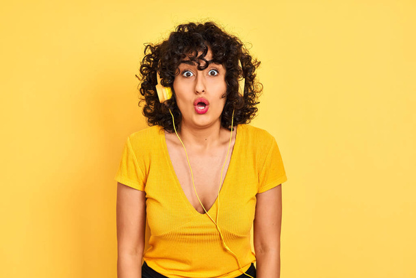 arabische Frau mit lockigem Haar, die Musik über Kopfhörer über isolierten gelben Hintergrund hört, ängstlich und schockiert mit überraschtem Ausdruck, Angst und aufgeregtem Gesicht. - Foto, Bild