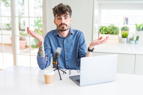 Young man recording podcast using microphone and laptop clueless and confused expression with arms and hands raised. Doubt concept. - Photo, Image