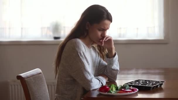 Girl sitting at table makes decision eat candies or vegetables - Filmati, video