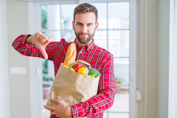 Όμορφος άνθρωπος κρατώντας τσάντα τρόφιμα με θυμωμένο πρόσωπο, αρνητικό σημάδι που δείχνει την αντιπάθεια με τους αντίχειρες κάτω, έννοια απόρριψη - Φωτογραφία, εικόνα