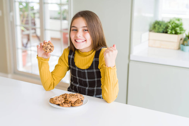 Belle jeune fille enfant manger des biscuits aux pépites de chocolat crier fier et célébrer la victoire et le succès très excité, acclamer l'émotion
 - Photo, image