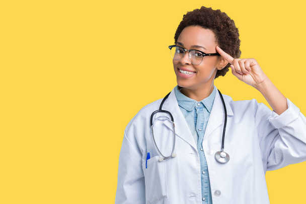 Joven mujer médica afroamericana con abrigo médico sobre fondo aislado Sonriendo señalando a la cabeza con un dedo, gran idea o pensamiento, buena memoria
 - Foto, Imagen