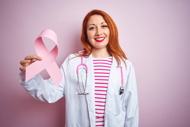 ピンクの孤立した背景の上に癌のリボンを持つ若い赤毛の医師の女性は、歯を示す自信のある笑顔で立って笑顔で立って、笑顔 - 写真・画像