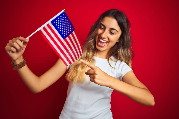 Kırmızı izole arka plan üzerinde Amerika bayrağı tutan genç kadın el ve parmak ile işaret çok mutlu - Fotoğraf, Görsel