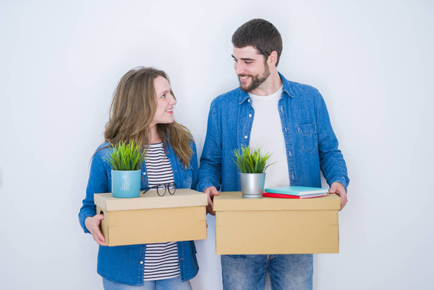 Jovem casal bonito segurando caixas de papelão muito feliz por se mudar para uma nova casa sobre fundo isolado branco
 - Foto, Imagem